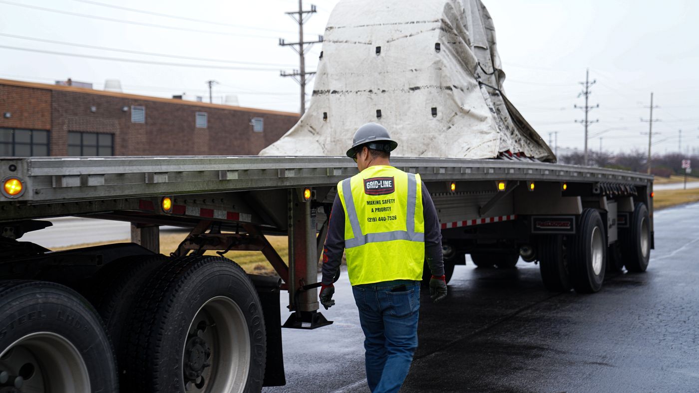 Grid-Line driver beside a tarped steel coil on a flatbed